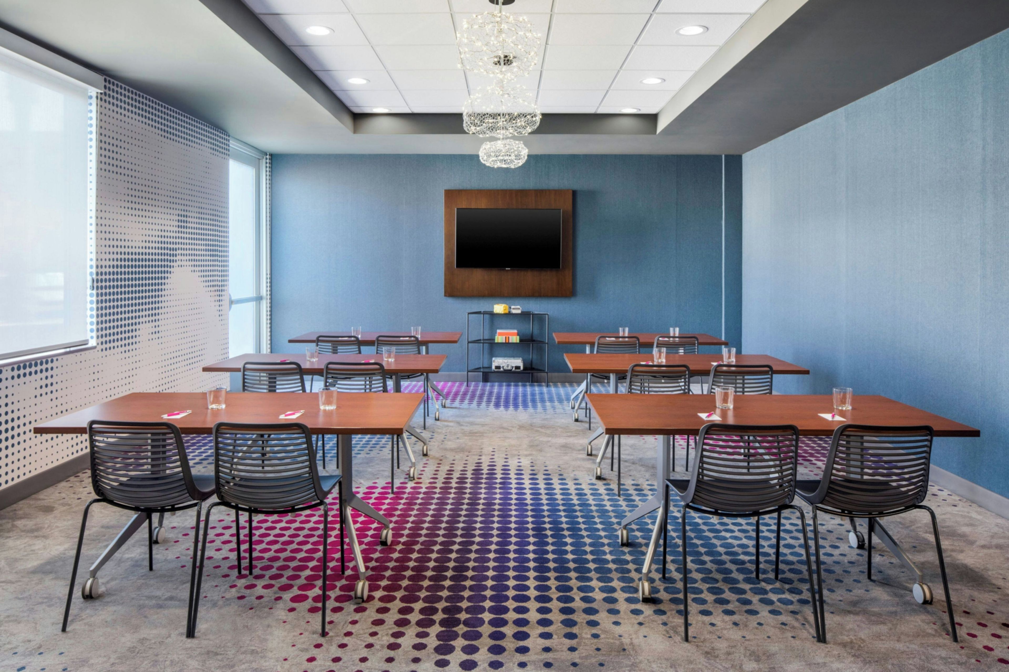 A conference room with a large flat-screen TV mounted on the wall, multiple tables with chairs, and a patterned carpet.