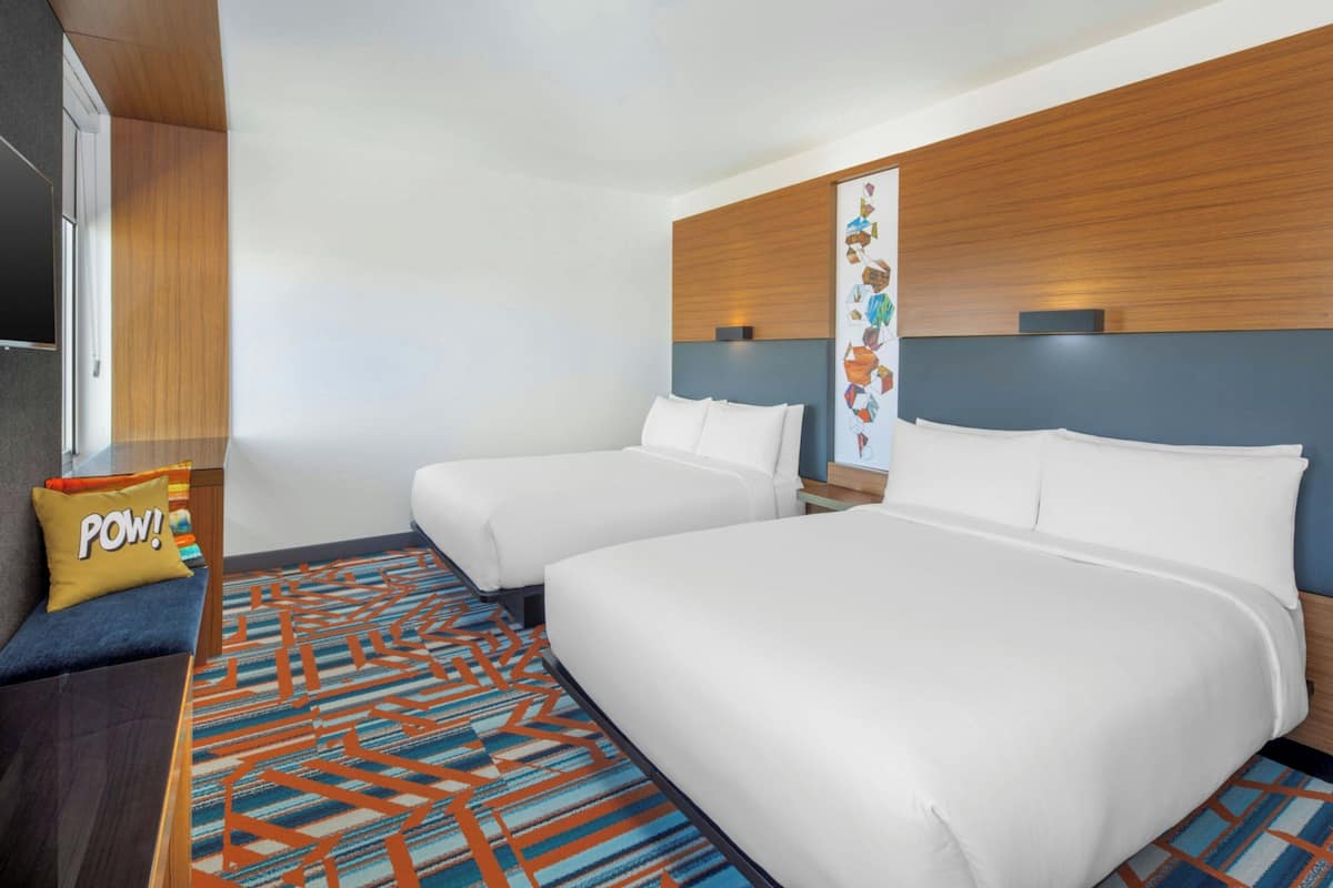 Habitación de hotel con dos camas, cabecero de madera, alfombra estampada y una obra de arte colgada en la pared.