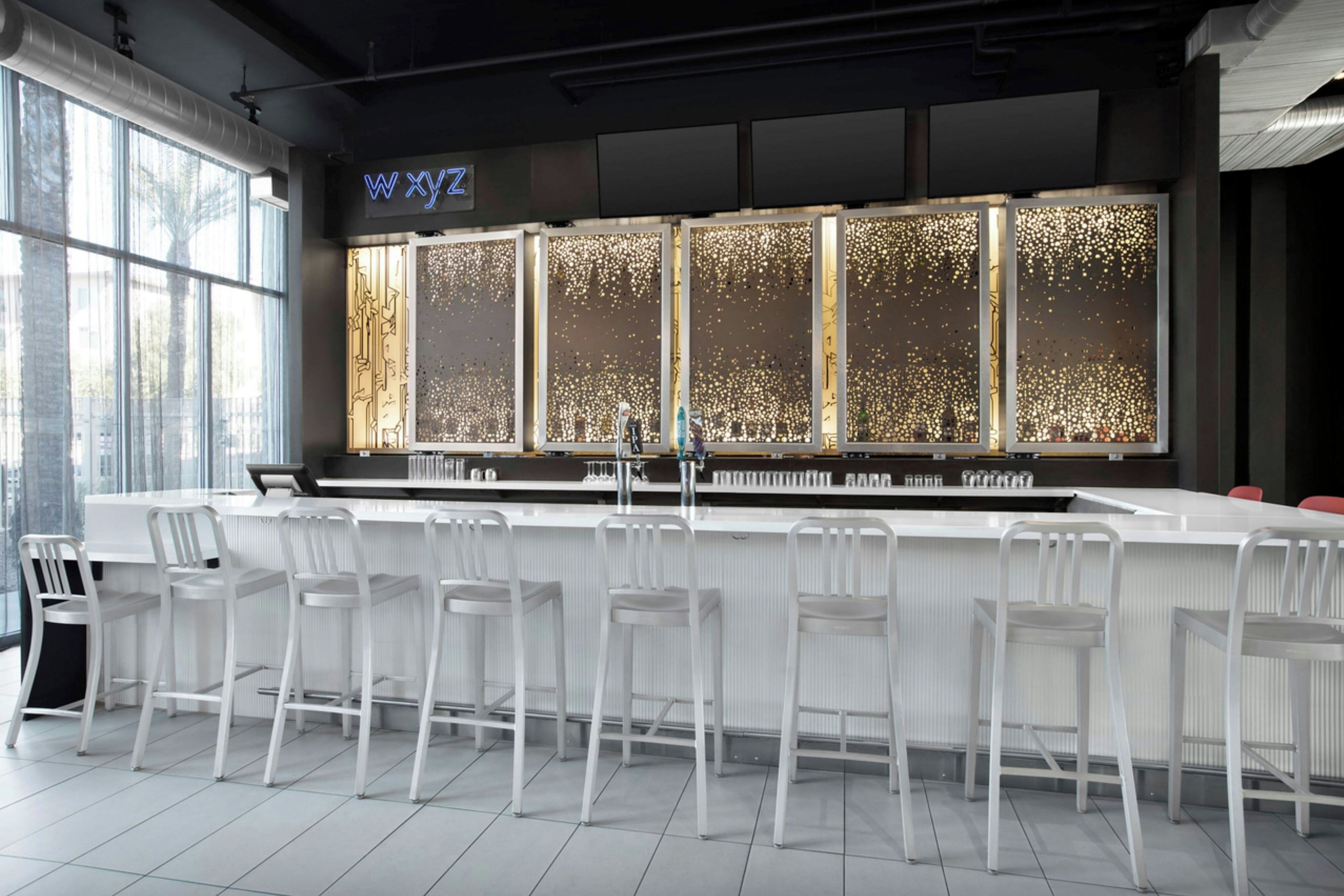 Eine moderne Bar mit einer langen Theke, weißen Barhocker und einem dekorativen Fenster mit einer Neonreklame.