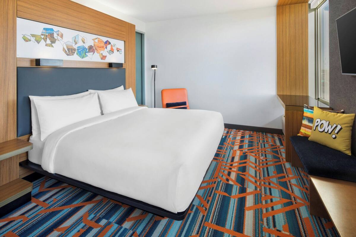 Habitación de hotel con una cama grande, un sofá con un cojín de colores, una alfombra estampada y una obra de arte colgada en la pared.