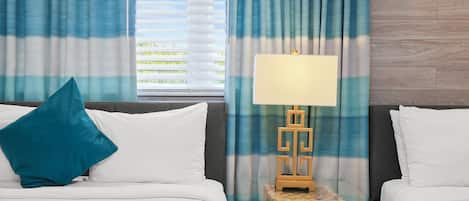 A hotel room with two beds, a nightstand with a lamp, and a window with blinds.