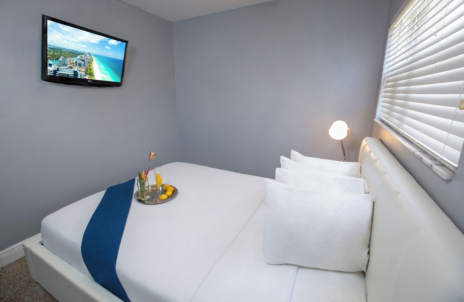 A hotel room with a bed, a TV on the wall, a bedside lamp, and a window with blinds.