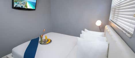 A hotel room with a bed, a TV on the wall, a bedside lamp, and a window with blinds.