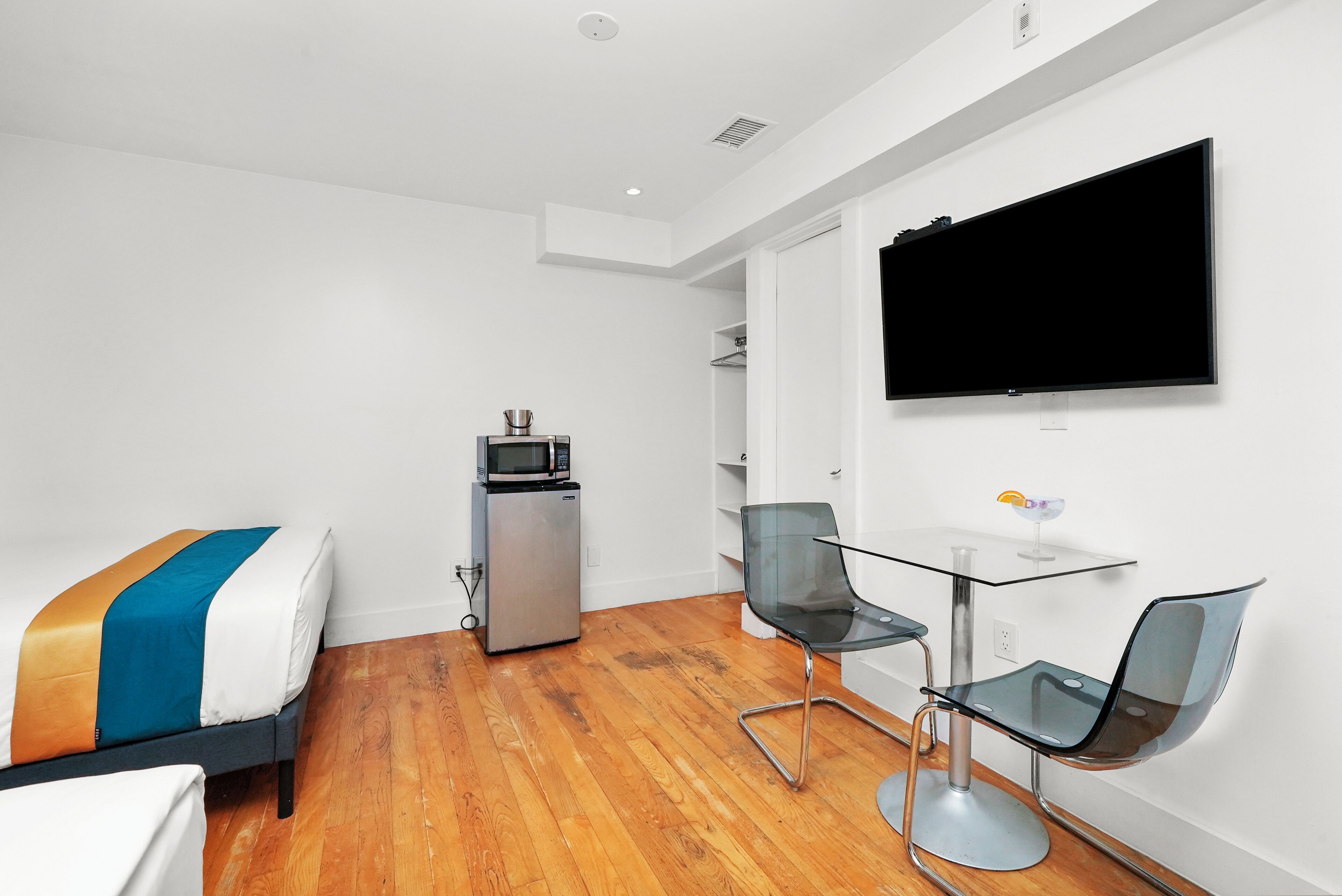 Habitació d'hotel moderna amb llit, petita cuineta, televisió penjada a la paret i taula de vidre amb dues cadires.