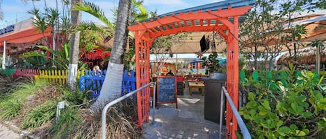 A covered outdoor seating area with a red archway, surrounded by greenery and a thatched-roof structure.