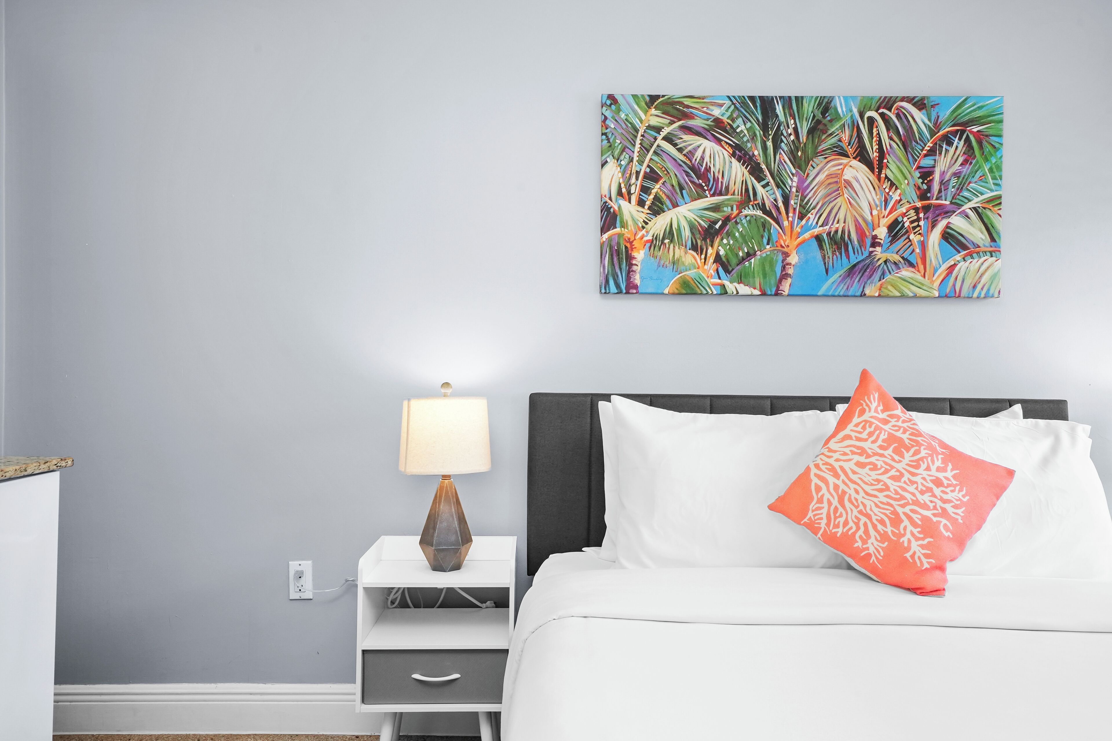 Un dormitorio con una cama, almohadas blancas, una mesita de noche con una lámpara y un cuadro en la pared con follaje tropical.