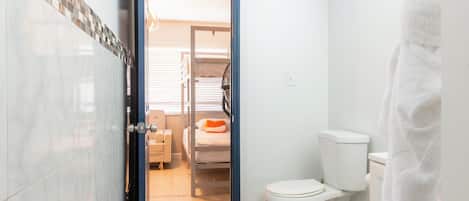 Un baño con inodoro blanco, una toalla colgada en la pared y una puerta que da a un dormitorio con literas y una silla.