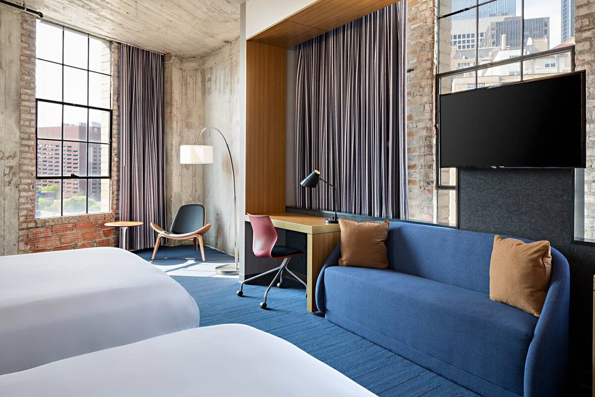 A hotel room with two beds, a desk, a chair, a sofa, a TV, and large windows with curtains.