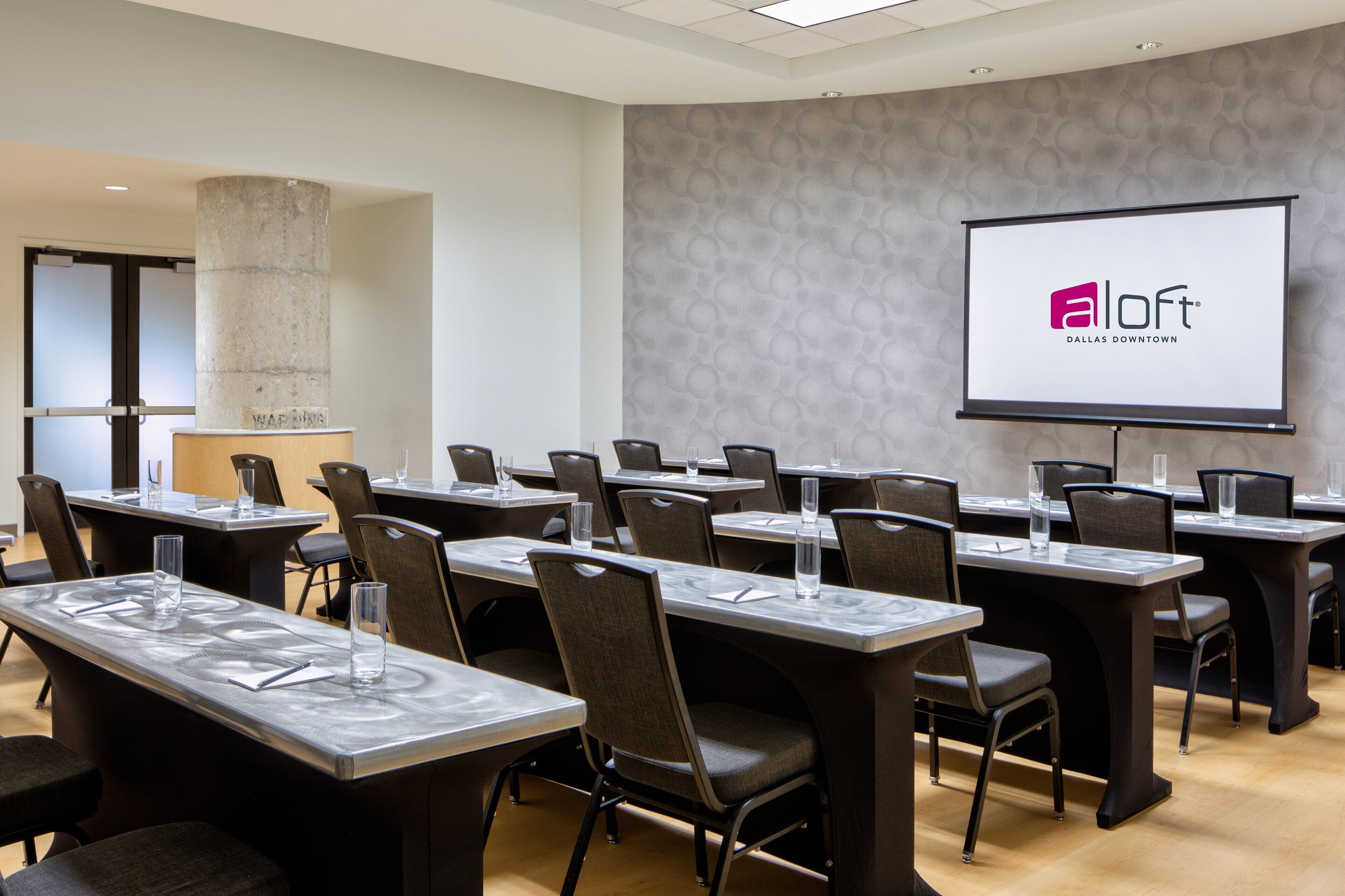 A conference room with multiple tables, chairs, and a projection screen displaying the logo "aloft Dallas Downtown".