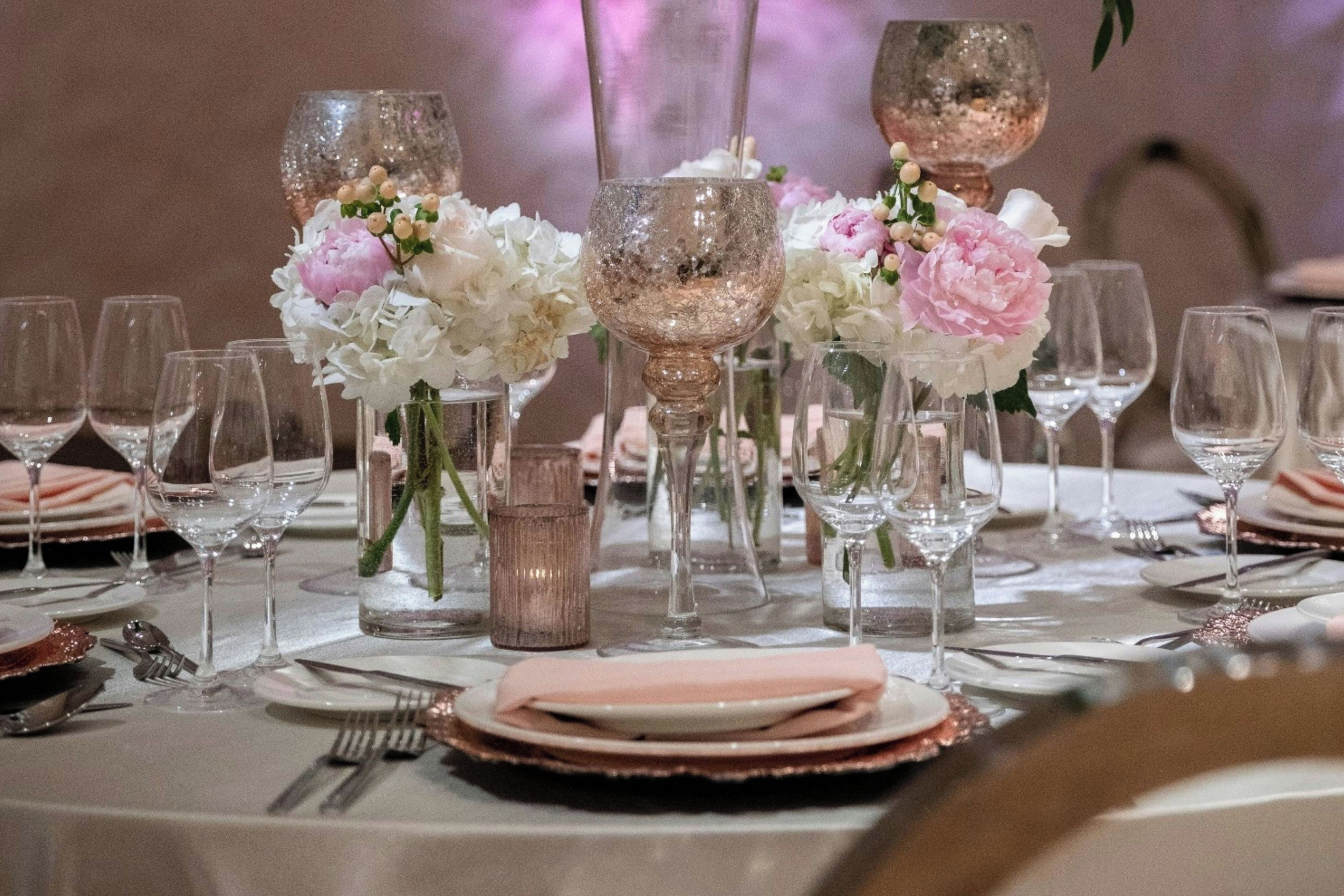 Una mesa dispuesta para un evento formal, con varias copas de vino, jarrones con flores y vajilla y cubiertos ordenados.