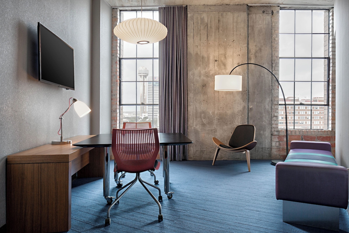 A hotel room with a desk, chair, bed, and large windows offering a city view.