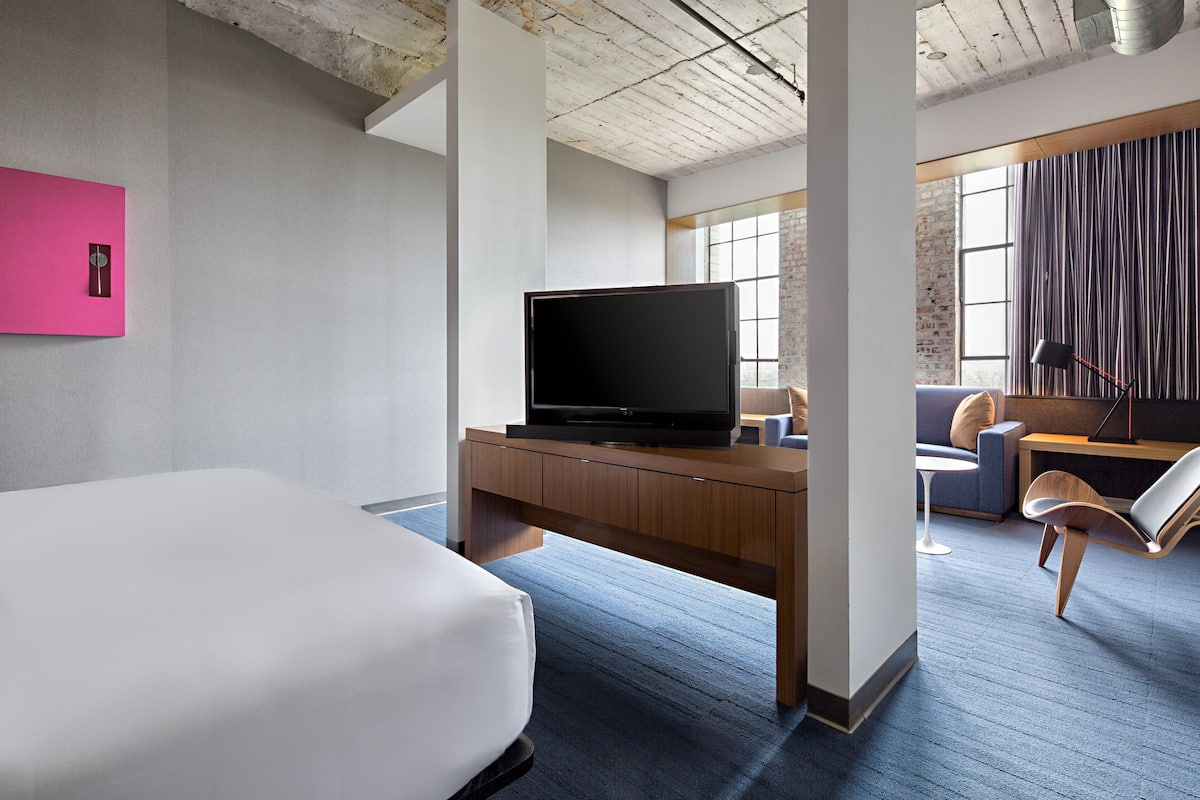 A modern hotel room with a large bed, a wooden desk with a flat-screen TV, a comfortable armchair, and a view of the city through large windows.