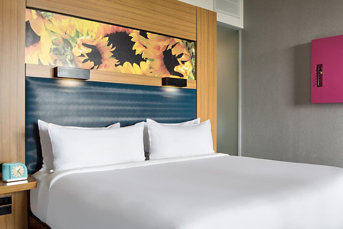 A hotel room with a bed, a nightstand with a clock, a wall-mounted TV, and a pink cabinet.