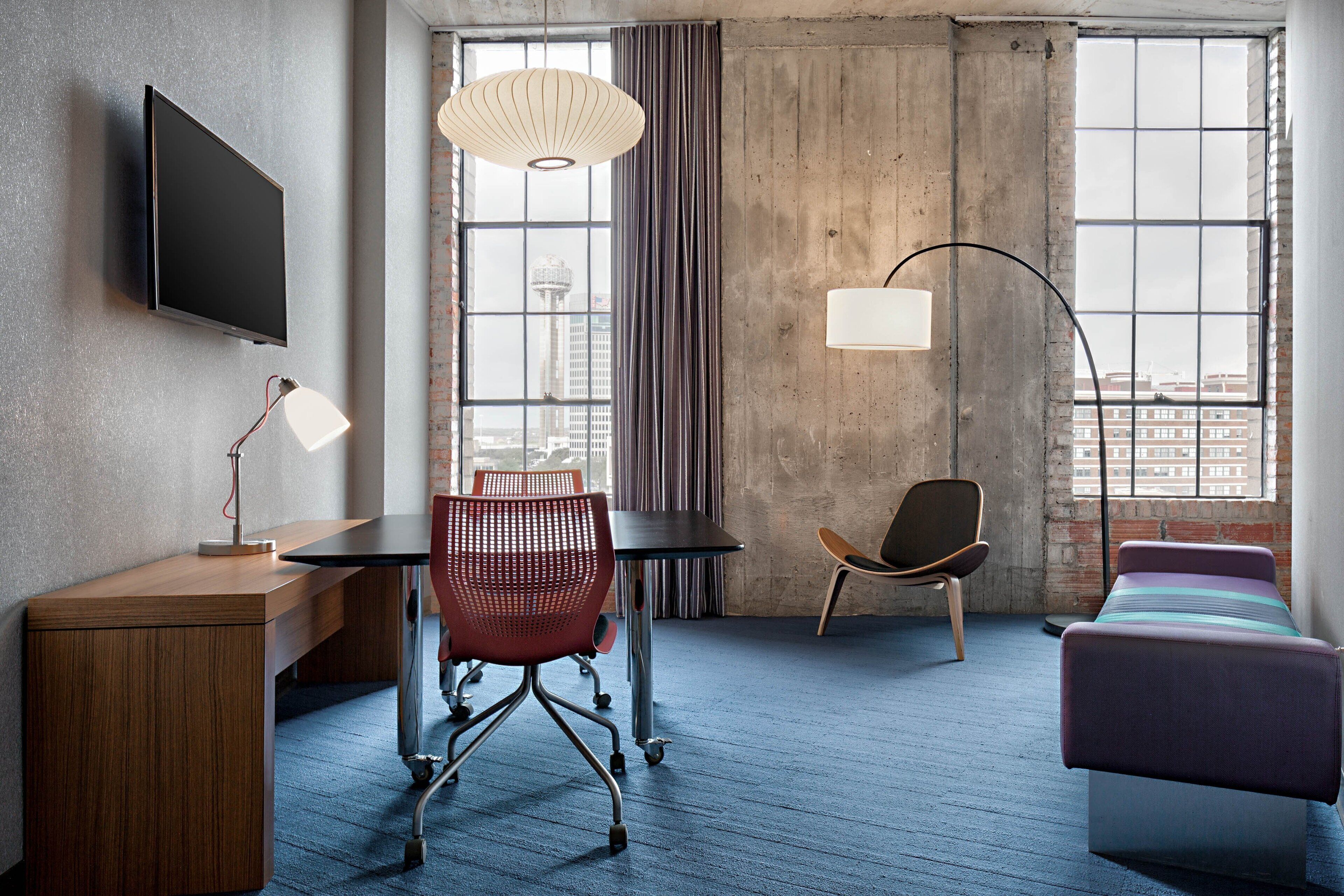 A hotel room with a desk, chair, bed, and large windows offering a city view.