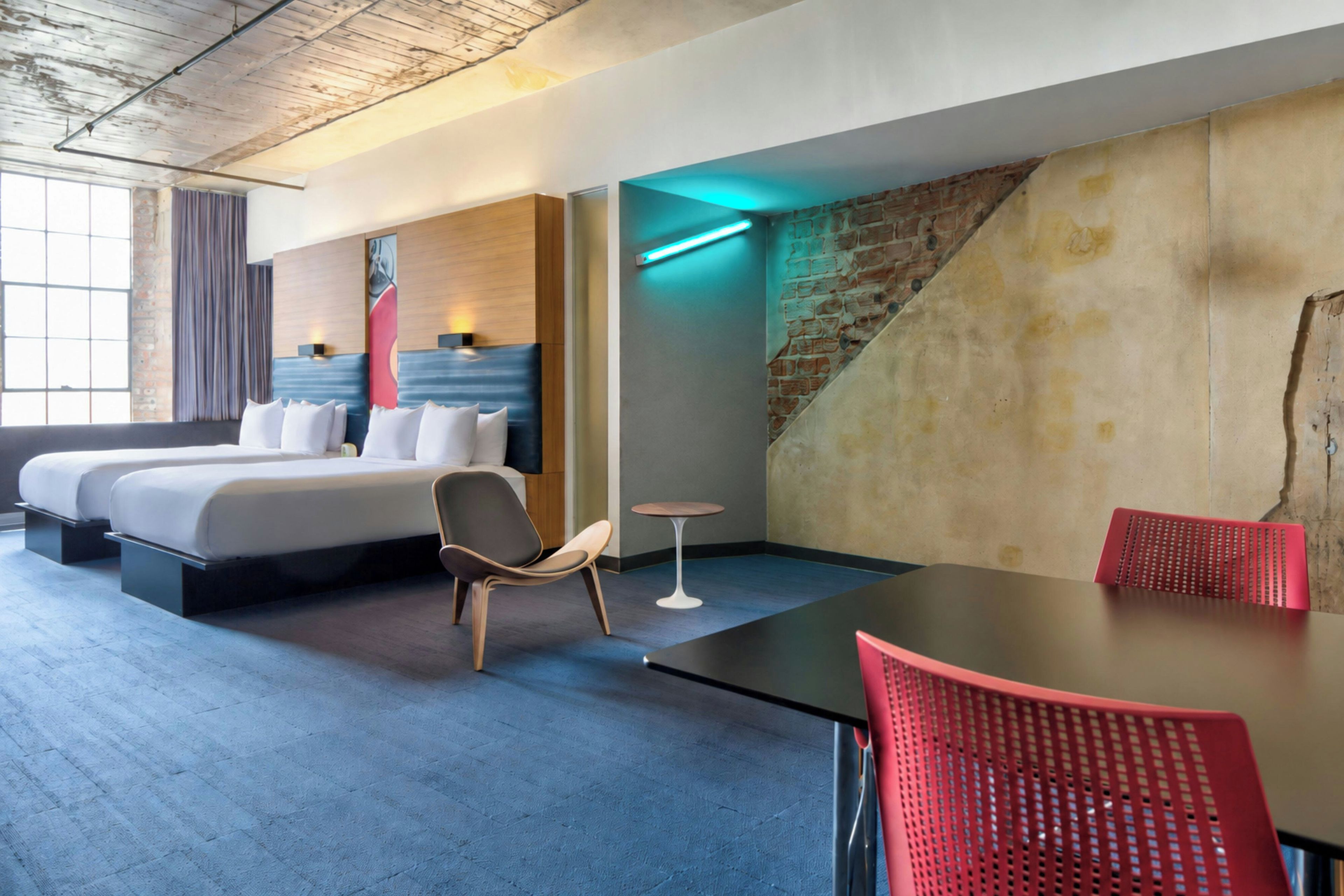 A hotel room with a bed, a chair, a table, and a brick wall.