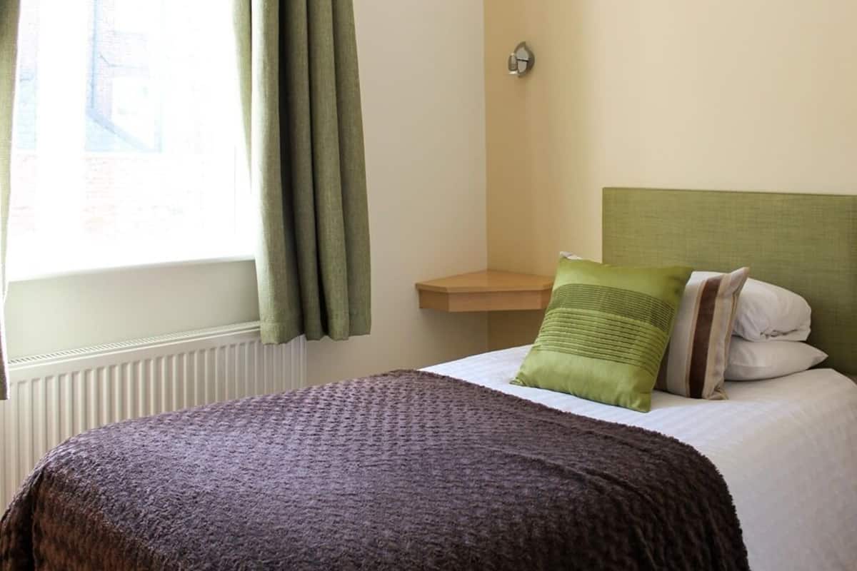 A hotel room with a bed, green curtains, a bedside table, and a wall-mounted light.