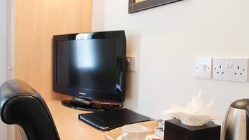 A hotel room with a wooden desk, a television, a cup, a tissue box, and a kettle.