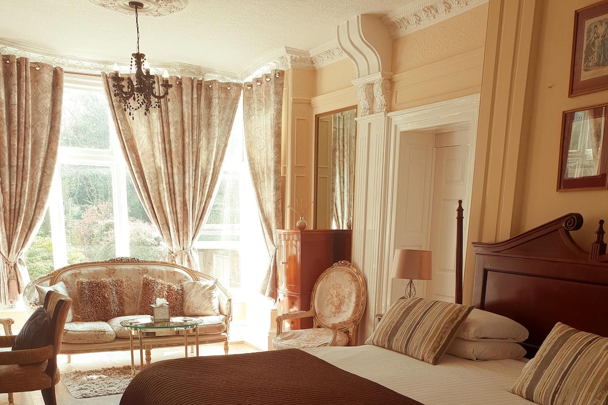 A well-lit bedroom with a large bed, a sitting area with a sofa and armchairs, and a view of the outdoors through a window.