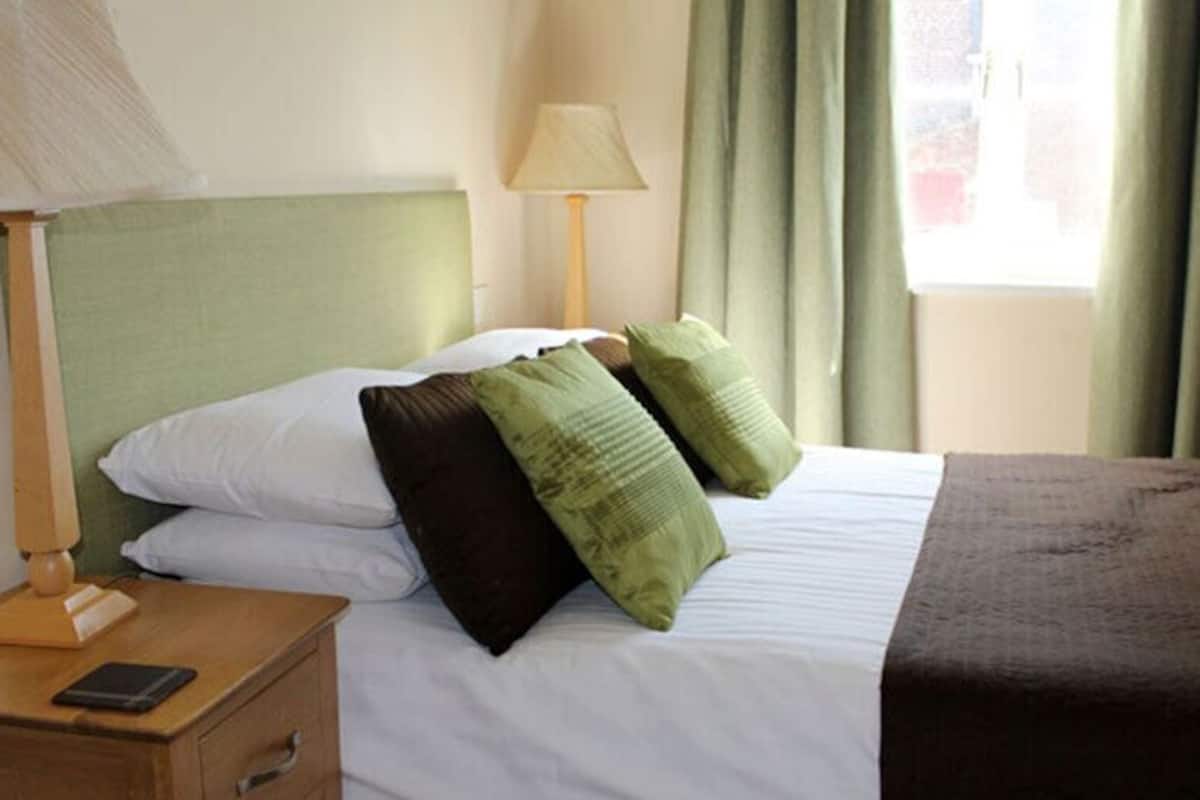 A neatly made bed with white and green pillows, a brown blanket, and a bedside table with a lamp and a smartphone.