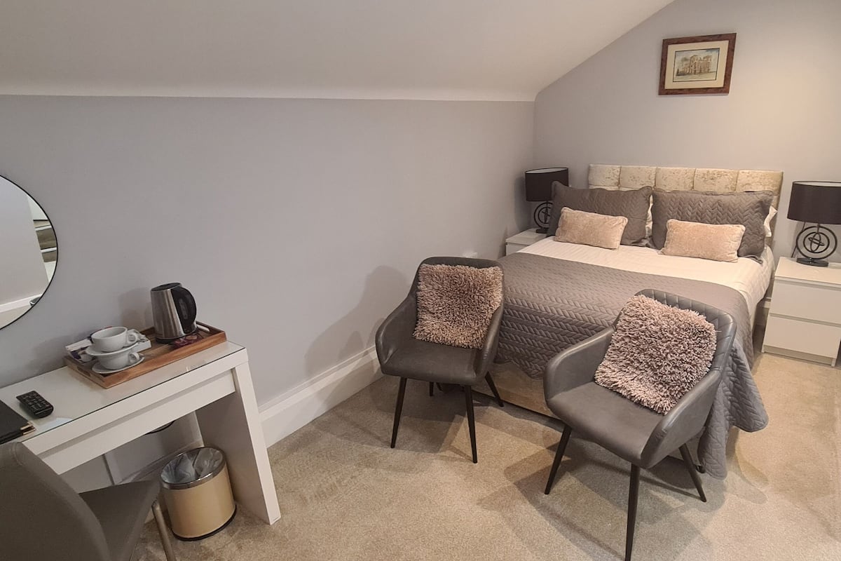 A bedroom with a bed, two chairs, a desk, a mirror, a radiator, and a bedside table with lamps.