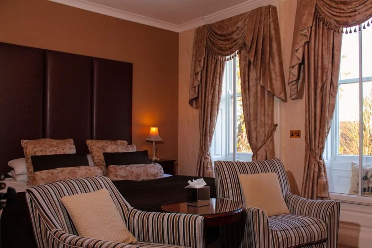 A hotel room with a bed, two striped armchairs, a small table, and a window with curtains.