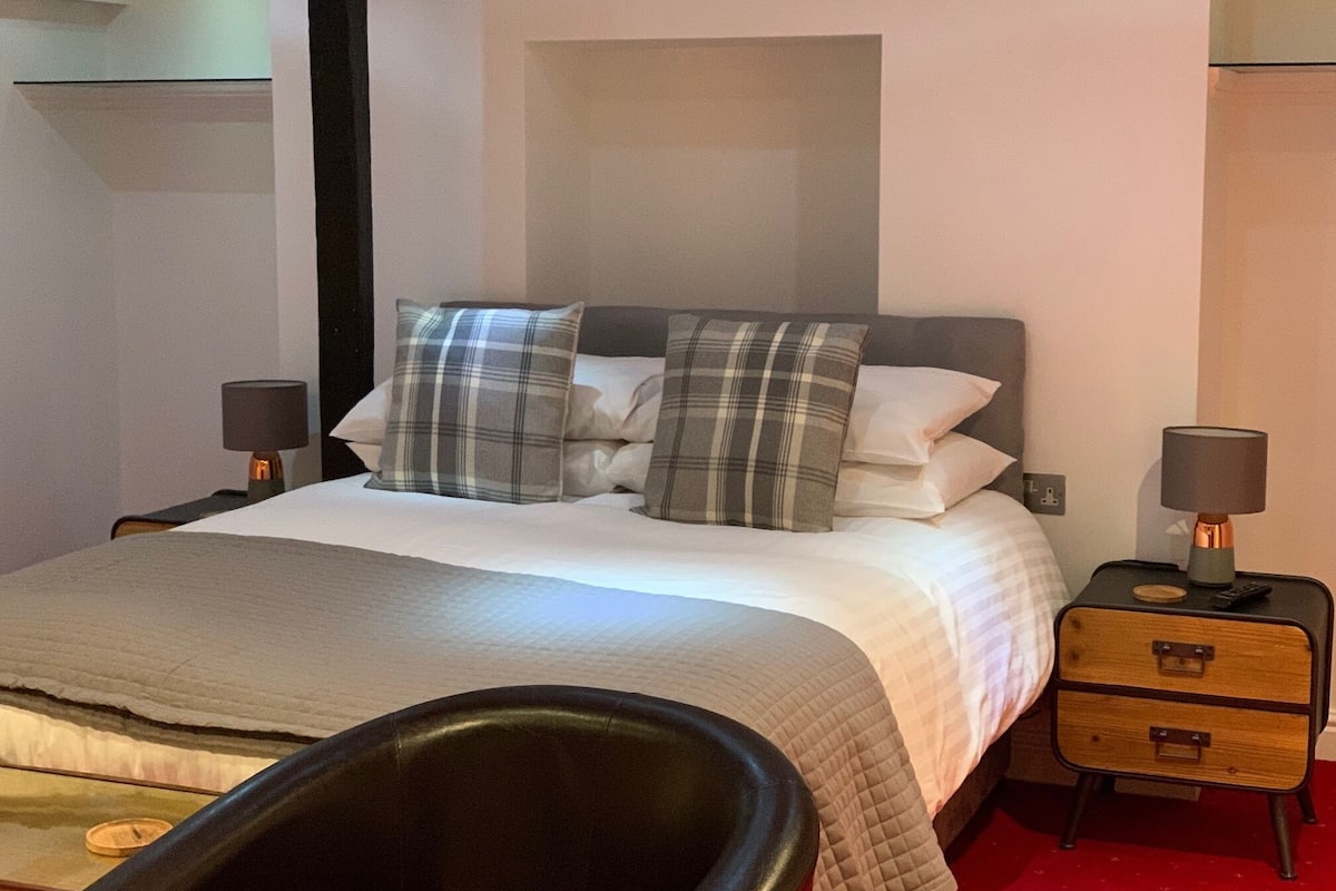 A neatly made bed with plaid pillows, a bedside table with a lamp, and a small wooden nightstand with drawers.
