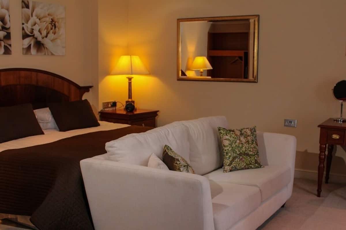 A hotel room with a bed, a white sofa, a nightstand with a lamp, a mirror, and a dresser.