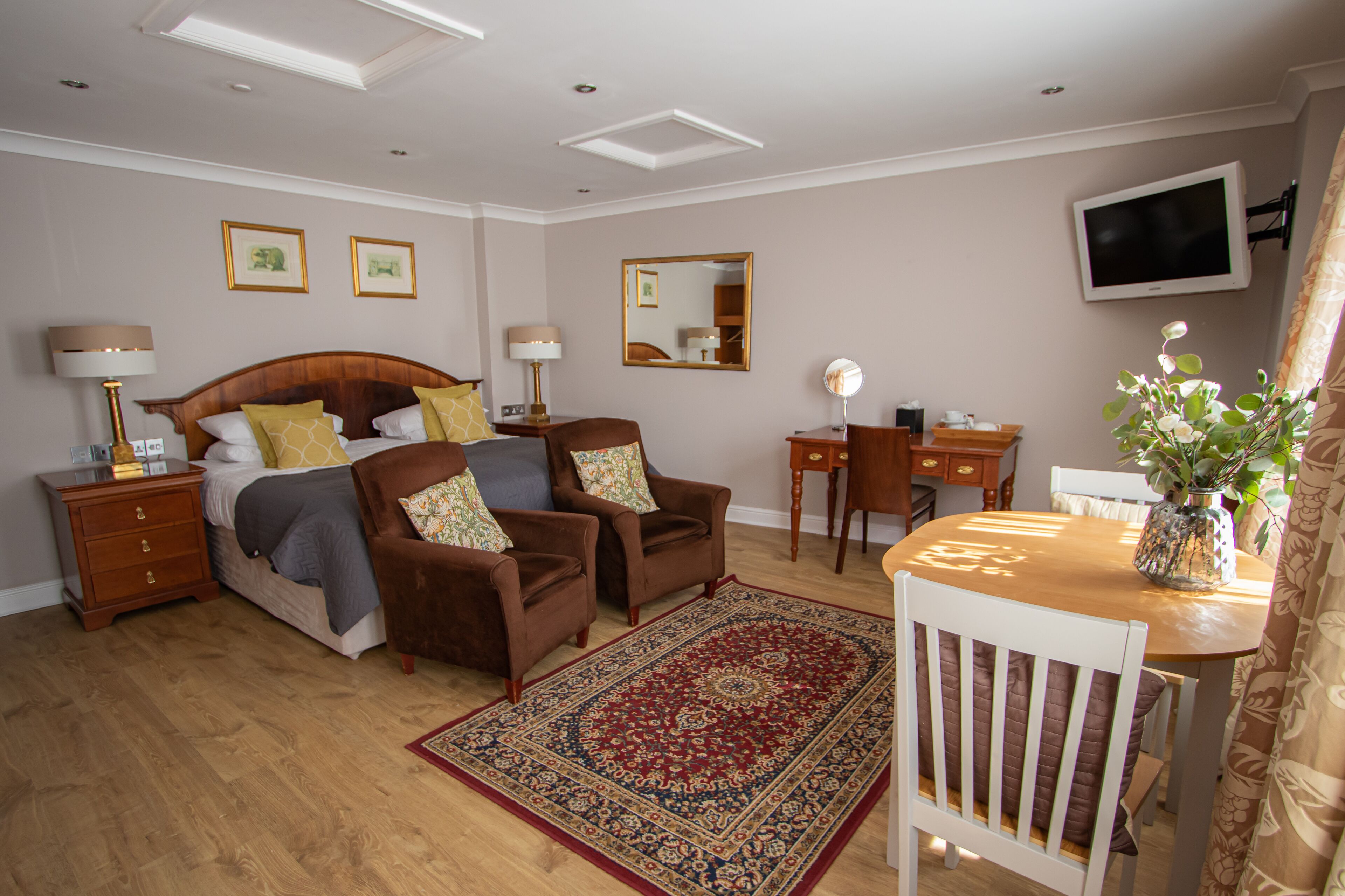A bedroom with a wooden bed, two brown armchairs, a wooden nightstand, a bedside table with lamps, a mirror, a TV, and a dining table with chairs.