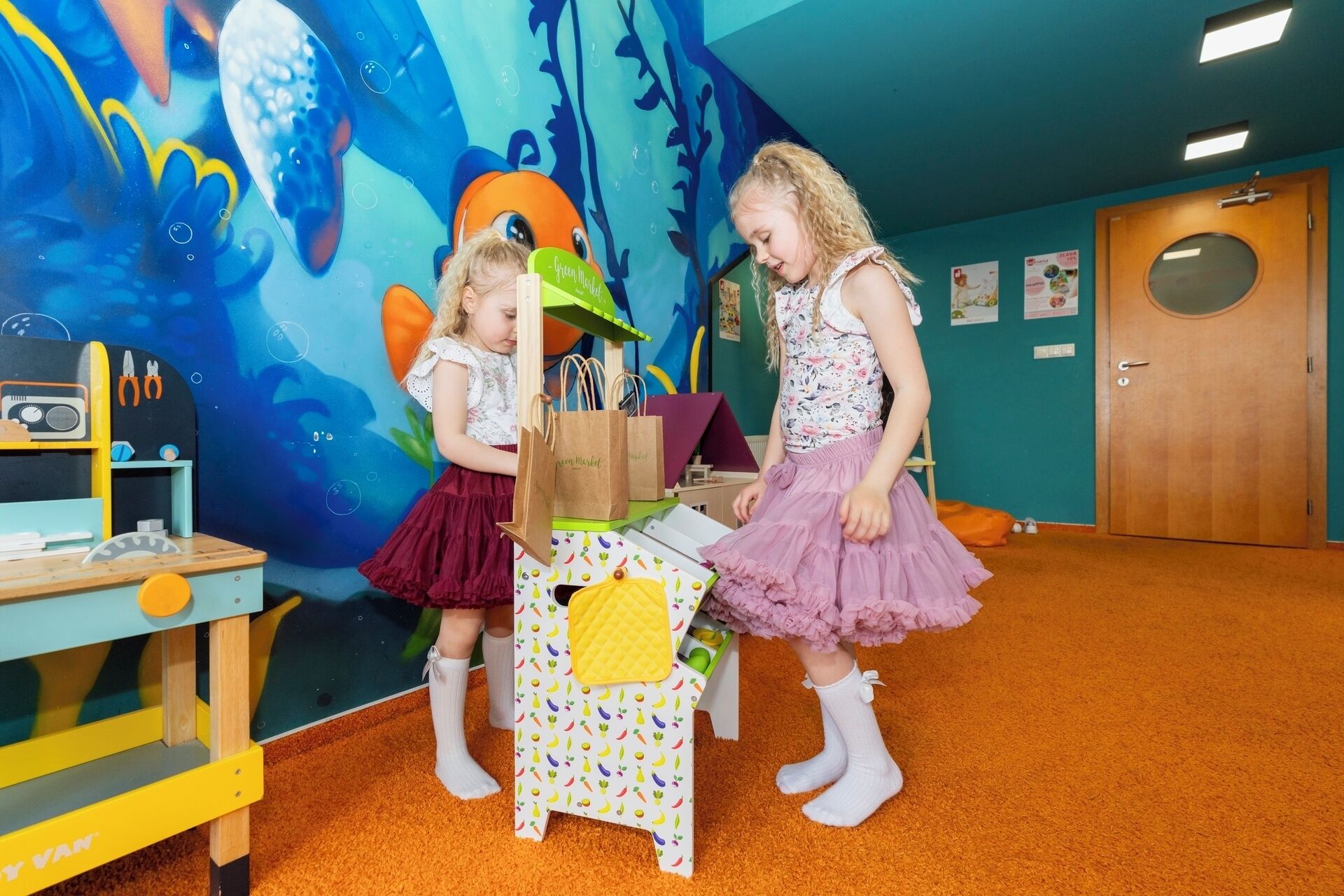 Two children in a room with a teal wall and orange carpet, a wooden table with a toy radio, and a door in the background.