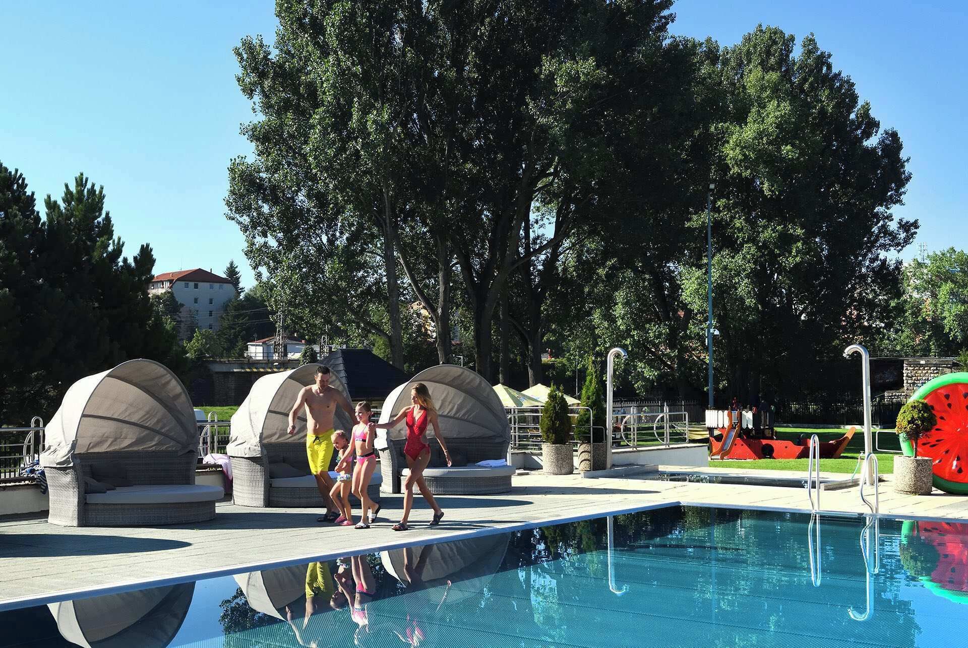 A swimming pool with lounge chairs and umbrellas, surrounded by trees and a building in the background.