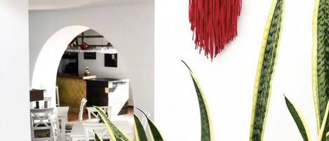 Un comedor con una mesa y sillas blancas, una decoración colgante de macramé y plantas grandes con rayas verdes y amarillas.