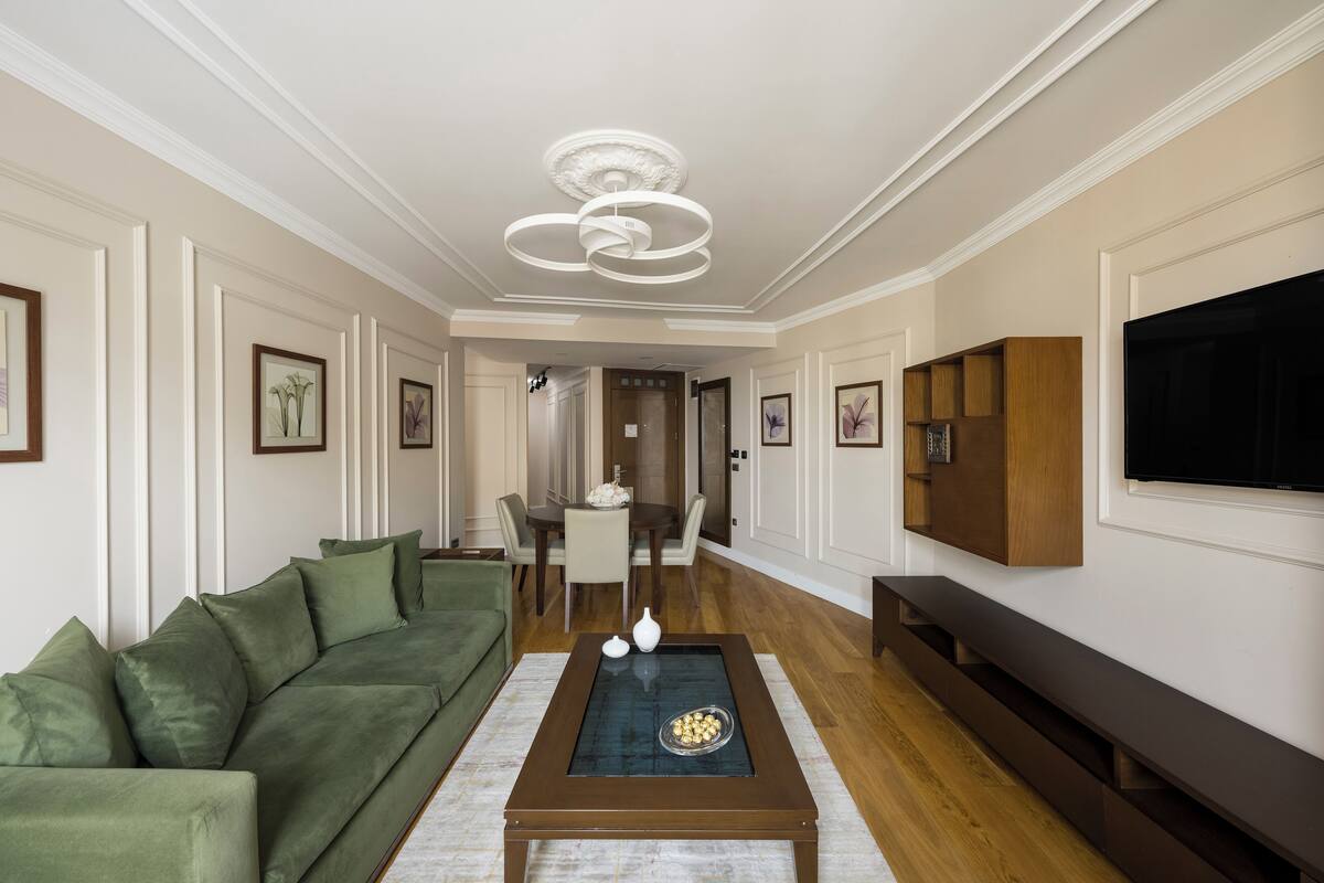 Un salon avec un canapé vert, une table basse, un téléviseur fixé au mur et, au fond, un coin repas.