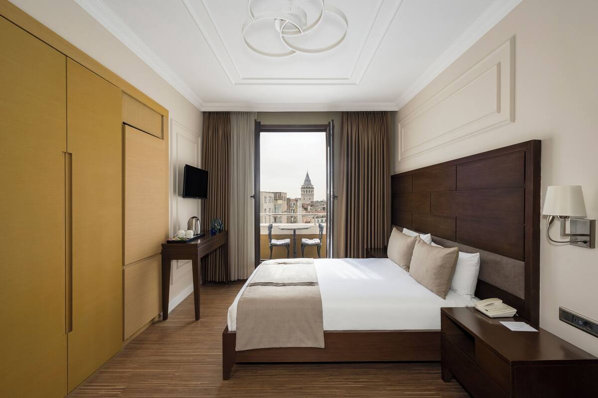 Une chambre d’hôtel avec un grand lit, une table de chevet avec une lampe, une télévision et une vue sur le paysage urbain par la fenêtre.