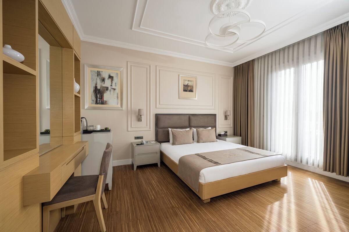 Une chambre d’hôtel comprenant un lit, des tables de chevet, un bureau, une chaise et une grande fenêtre avec des rideaux.