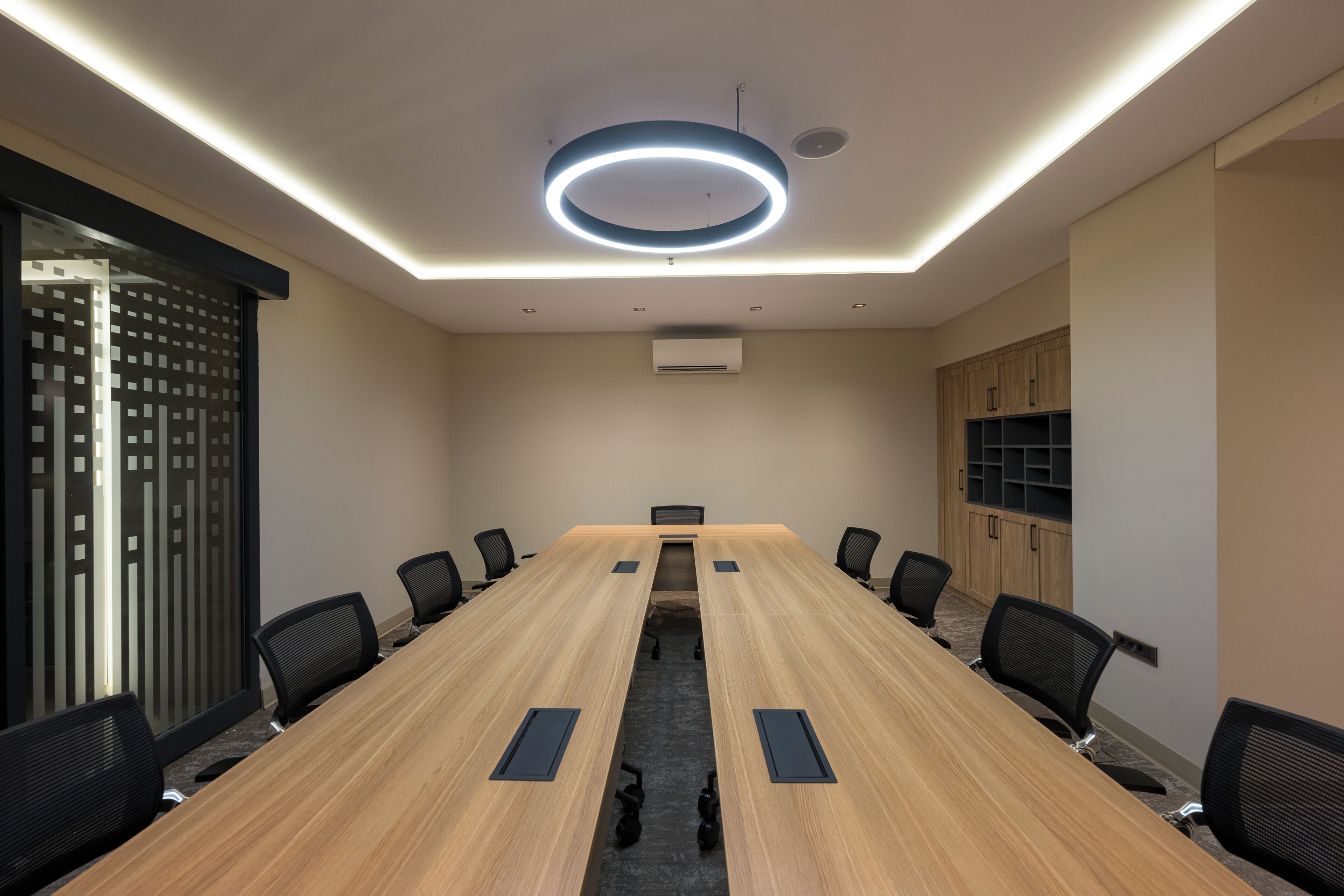 Een vergaderruimte met een lange houten tafel, zwarte stoelen en een moderne plafondlamp.