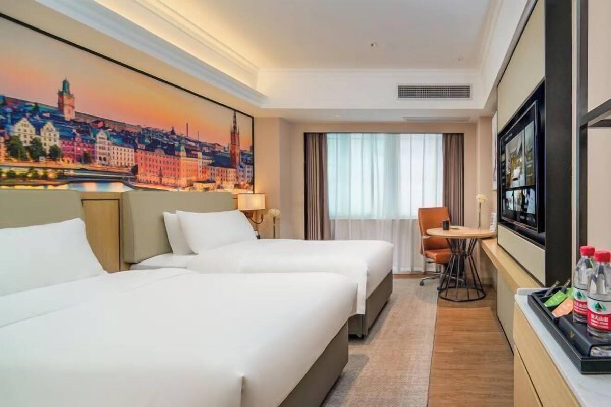 A hotel room with two beds, a desk, a chair, a TV, and a painting of a cityscape.
