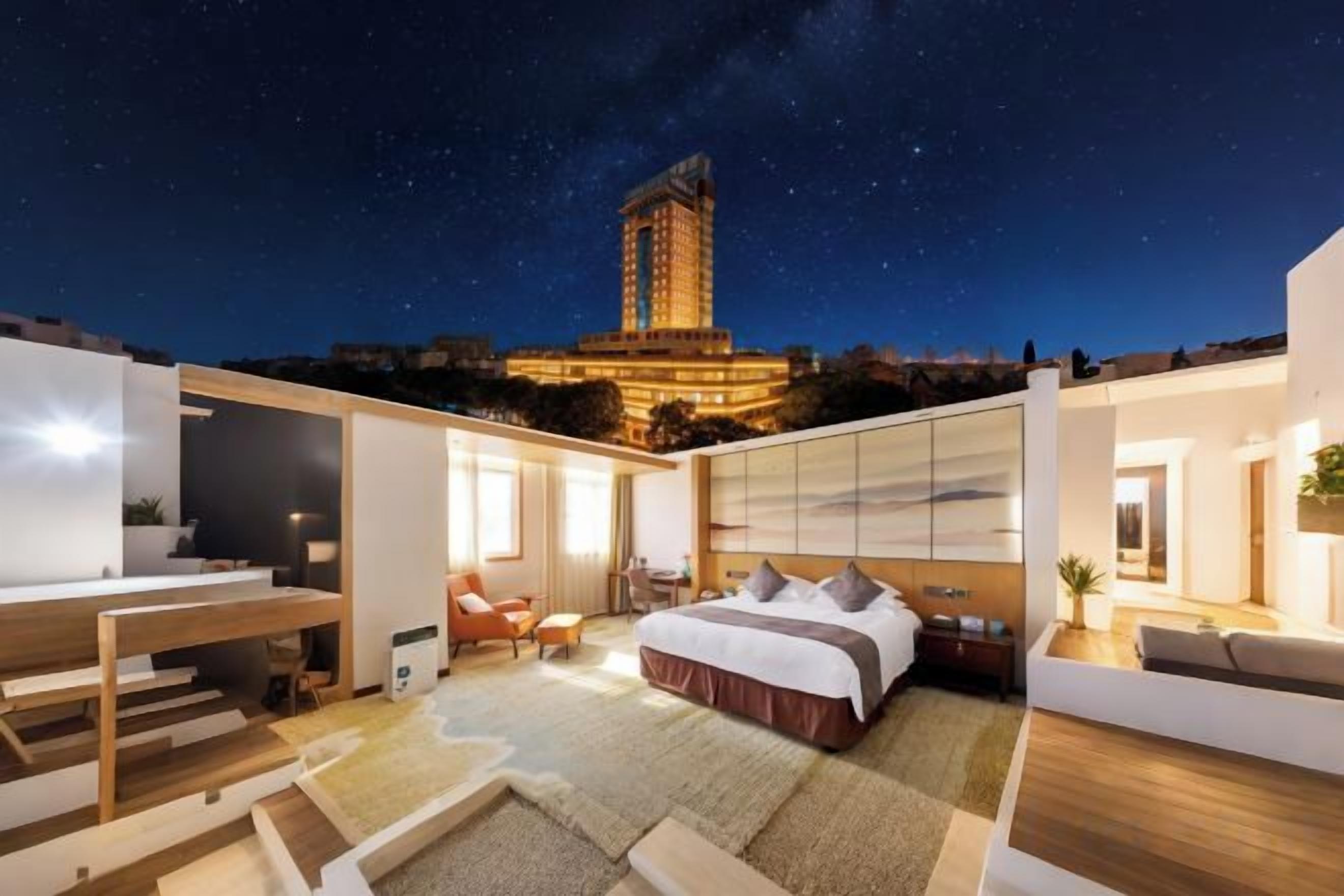 A modern hotel room with a large bed, a desk, and a view of a cityscape at night.