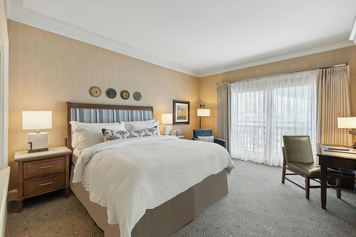 A hotel room with a large bed, a desk, a chair, a nightstand, and a window with curtains.