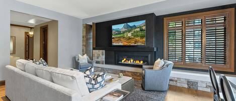 Una sala moderna con un televisor de pantalla plana grande montado sobre una chimenea, un sofá gris con cojines estampados y un comedor con mesa y sillas de madera.