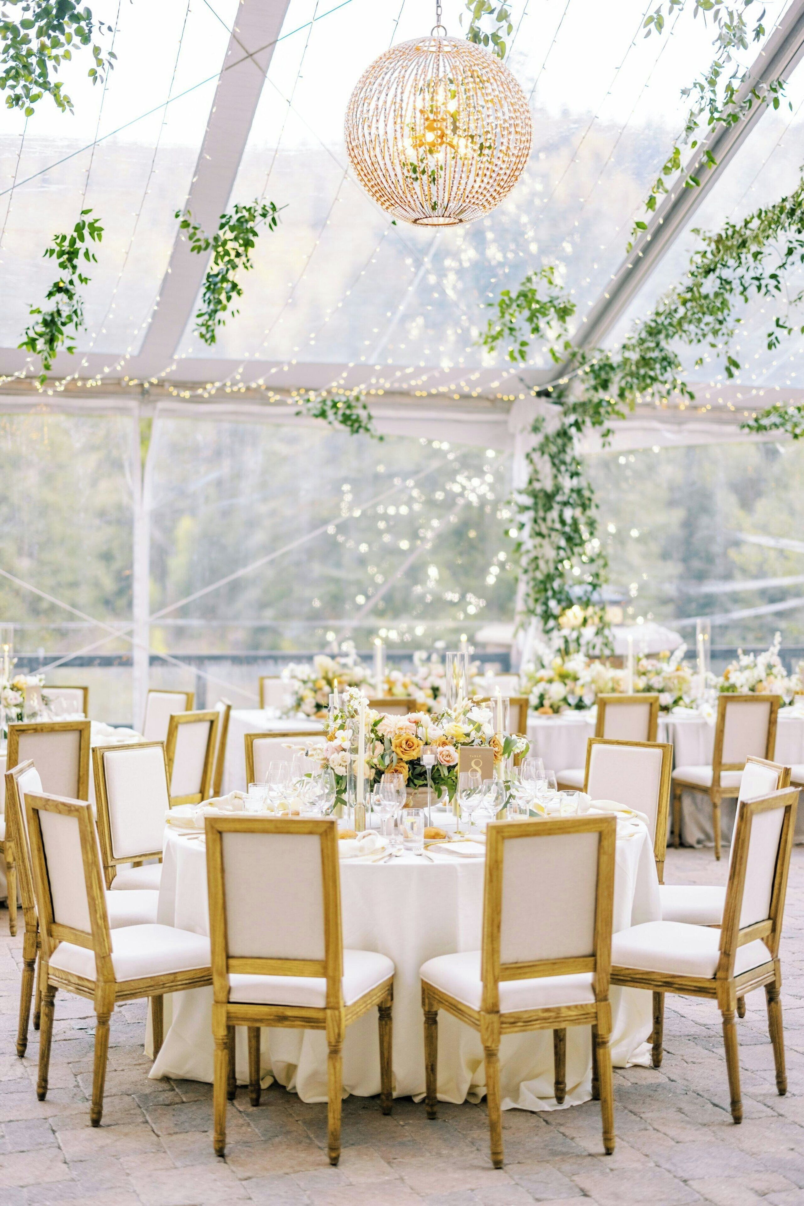 A tented outdoor event space with round tables, white tablecloths, and gold chairs.