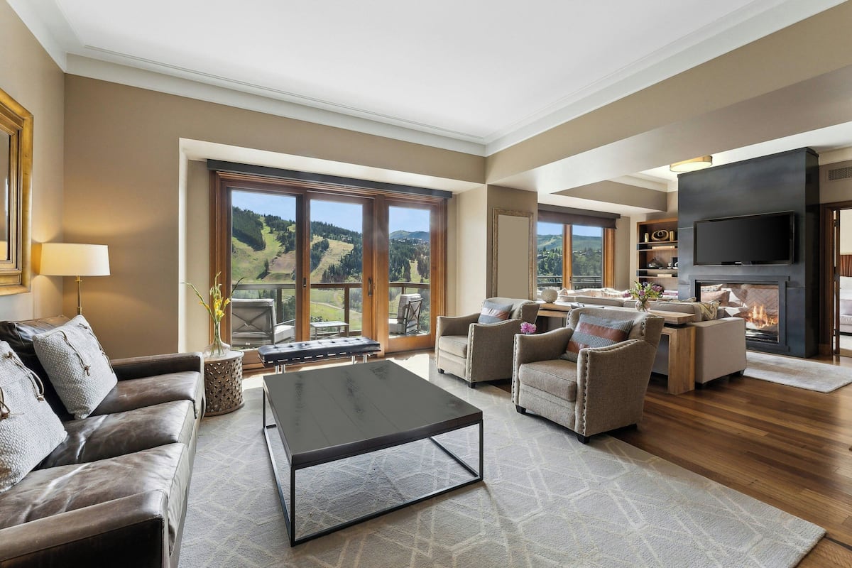 A spacious living room with a fireplace, large windows offering a view of the countryside, and a flat-screen TV mounted on the wall.