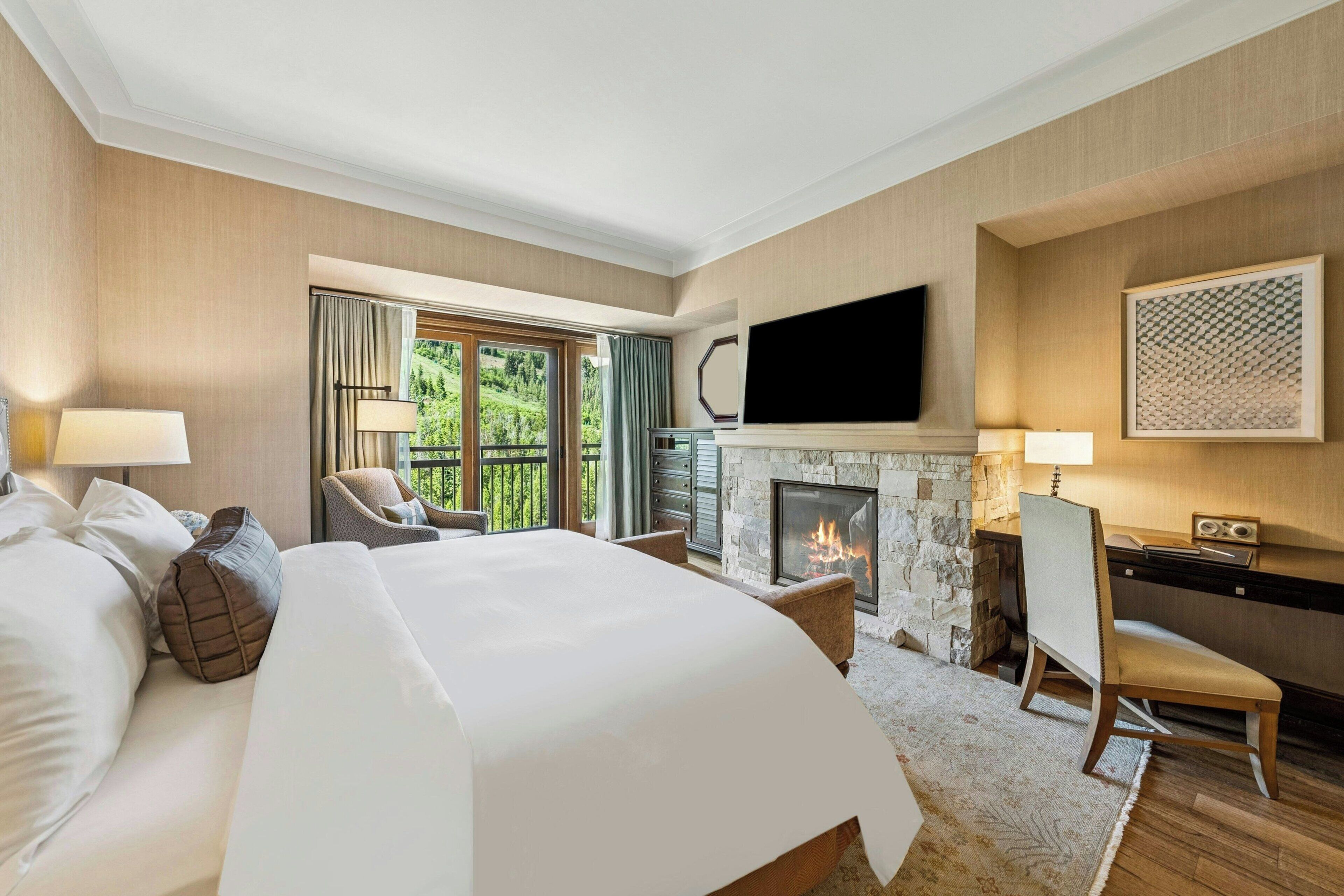 A hotel room with a large bed, a fireplace, a television, and a balcony with a view of greenery.