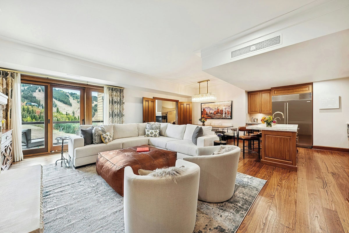 A spacious living room with a large sectional sofa, a wooden coffee table, and a view of a mountainous landscape through large sliding glass doors.