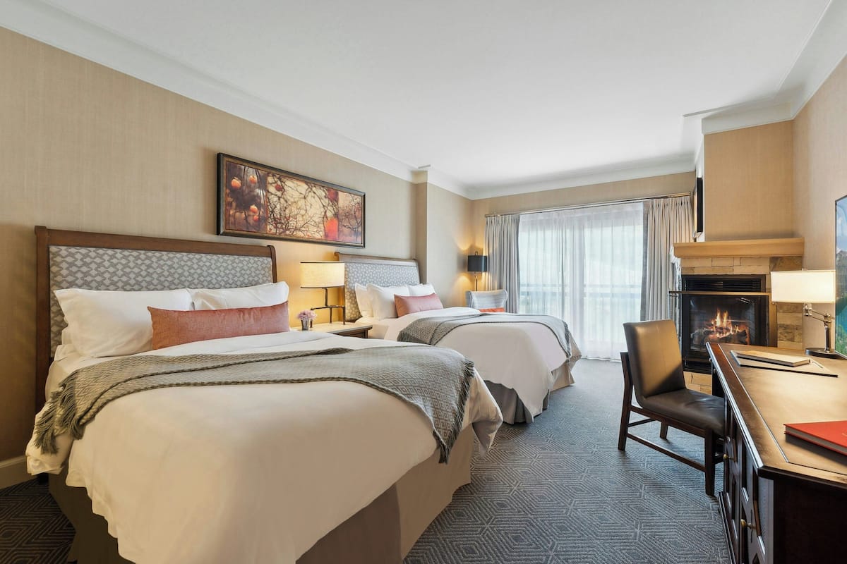 A hotel room with two beds, a desk, a chair, a fireplace, and a large window with curtains.