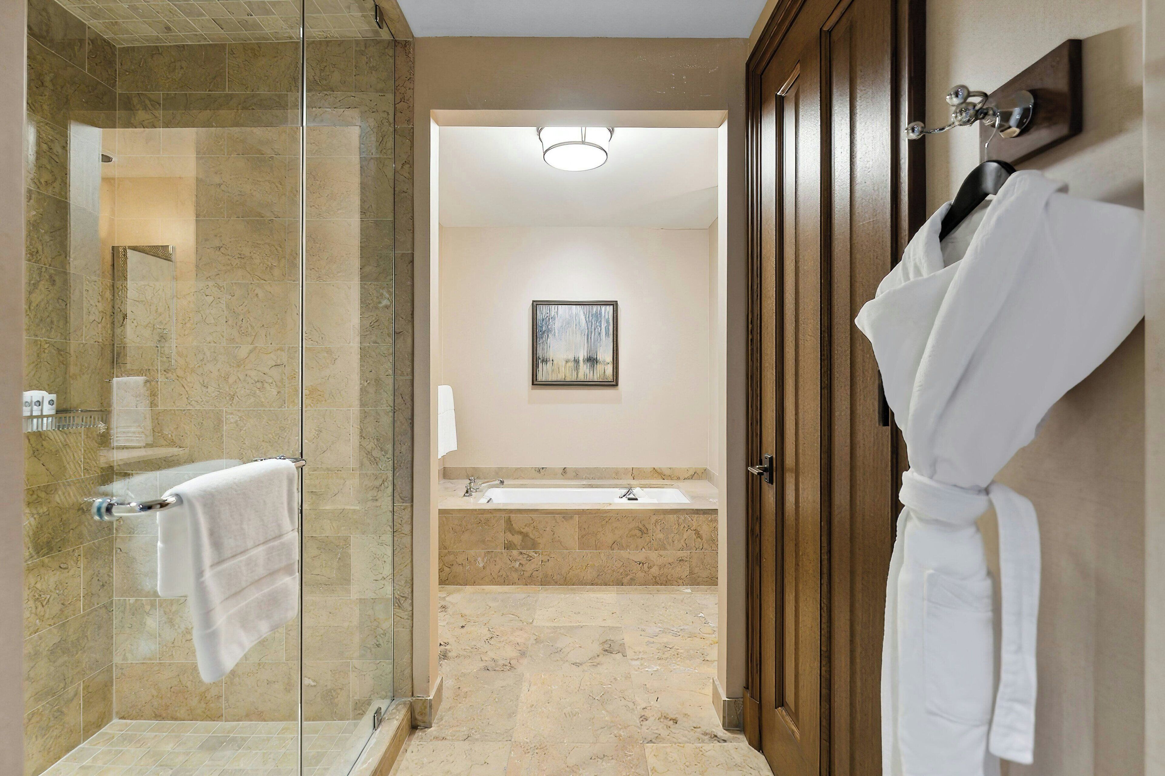 A modern bathroom with a glass-enclosed shower, a large bathtub, and a wooden door with a robe hanging on it.