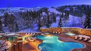 Piscina aquecida ao ar livre com espreguiçadeiras e uma parede de pedra, cercada por árvores cobertas de neve e um cenário montanhoso.