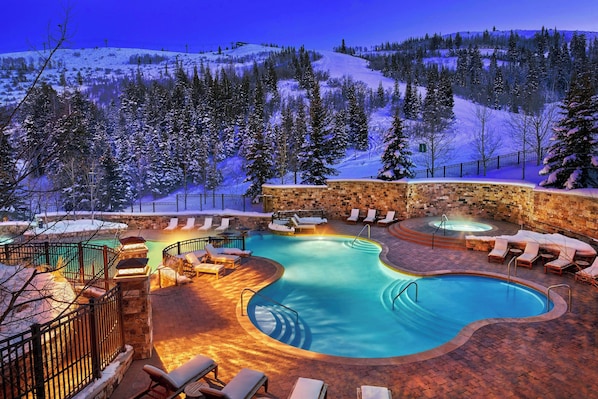 Una piscina climatizada al aire libre con sillones de descanso y un muro de piedra, rodeada de árboles cubiertos de nieve y un paisaje montañoso.