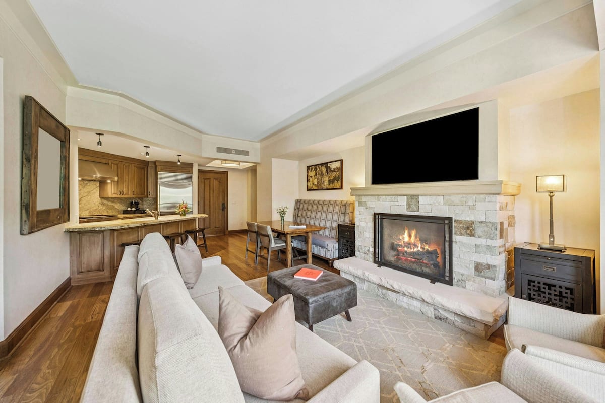 A living room with a fireplace, a television mounted above it, and a dining area with wooden cabinets and a kitchen in the background.