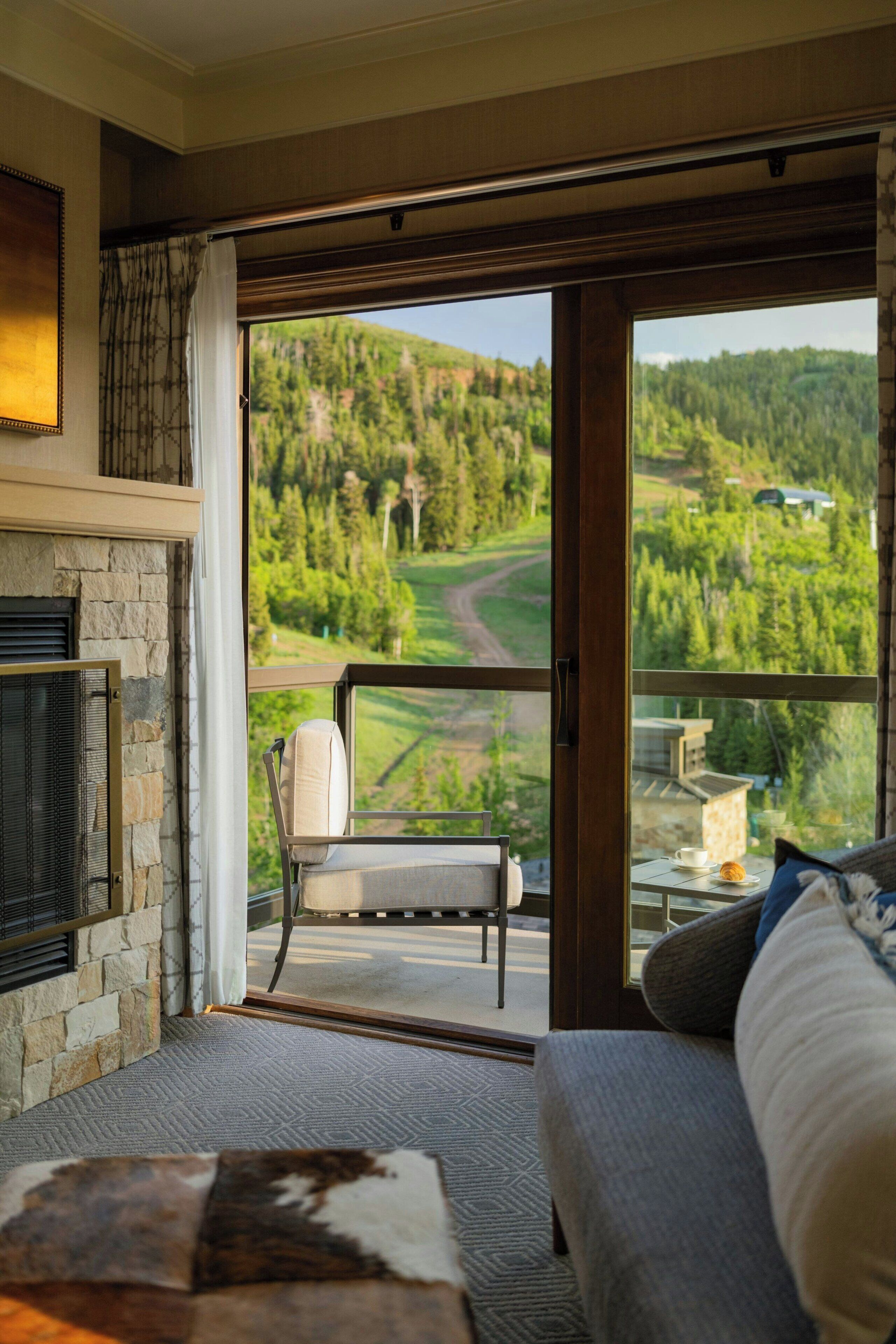 Ein Zimmer mit Kamin aus Stein, einer Couch, einem Sessel und Blick auf eine grüne Landschaft durch eine Glastür.