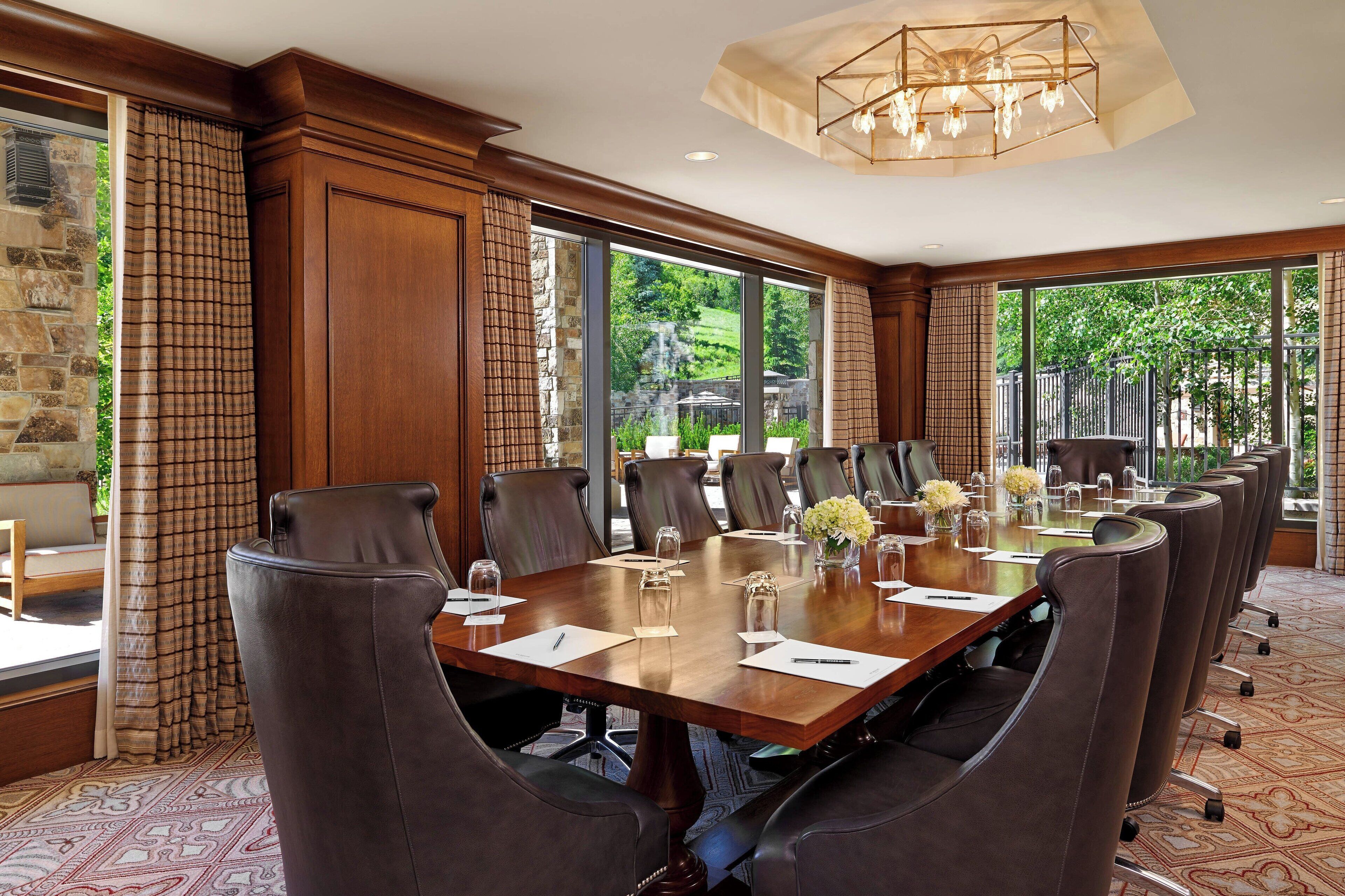 A conference room with a large wooden table, leather chairs, and large windows offering a view of greenery outside.